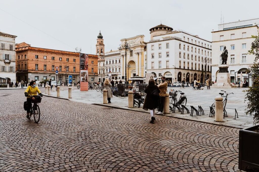 A woman in a yellow jacket and wearing a face mask bikes her way across Parma's main square
