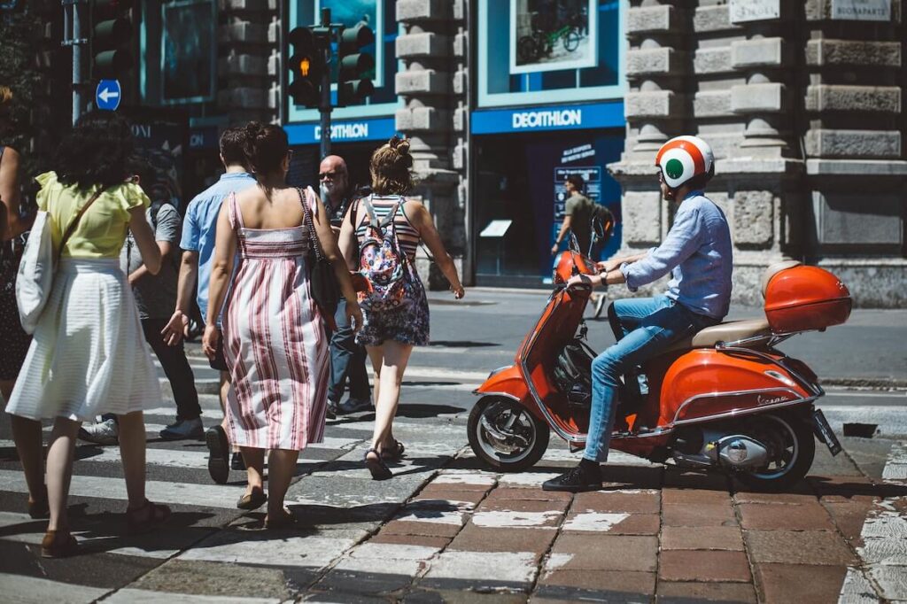 A young man on a scooter passes tourists at a pedestrian crossing in Milan