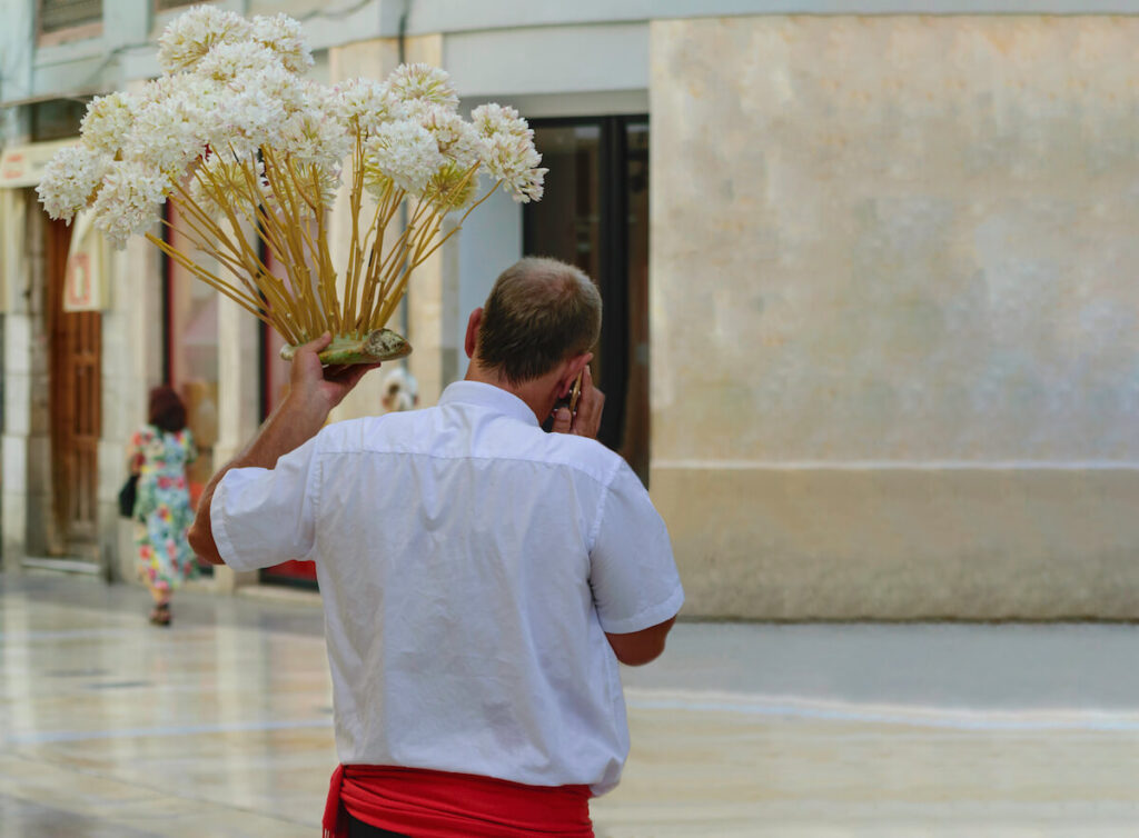 Rear view of a a "Biznaguero", man wearing black trousers, red waistband and white shirt who sells a handcrafted "flower" made of Jasmine called Biznaga