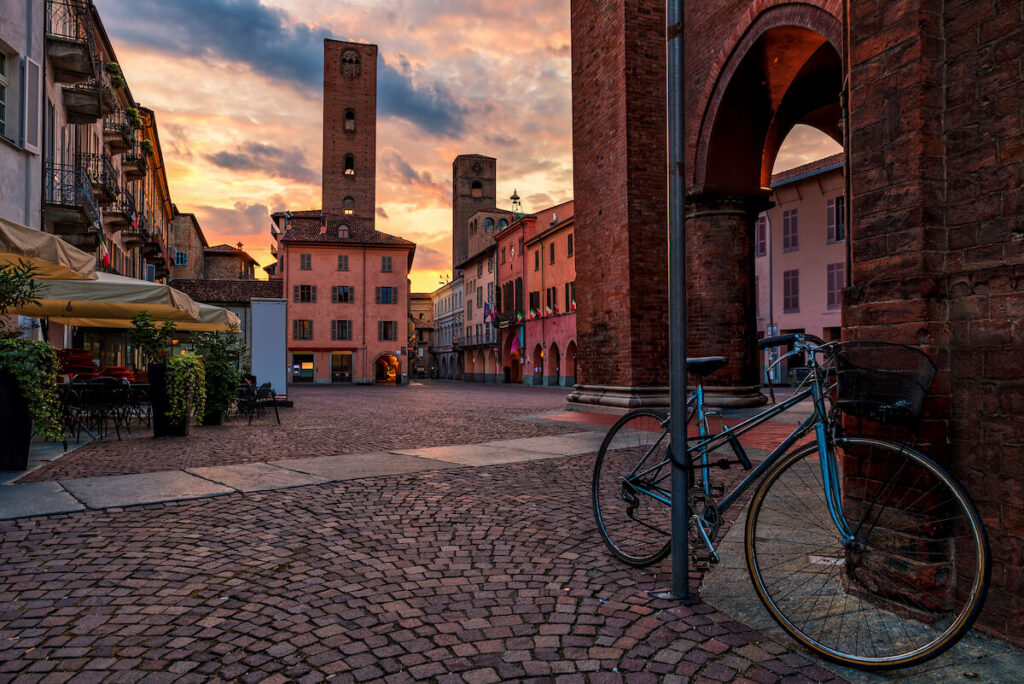 Bicycle on cobblestone town square surrounded by old houses and medieval towers under beautiful evening sky in Alba, Piedmont, Northern Italy