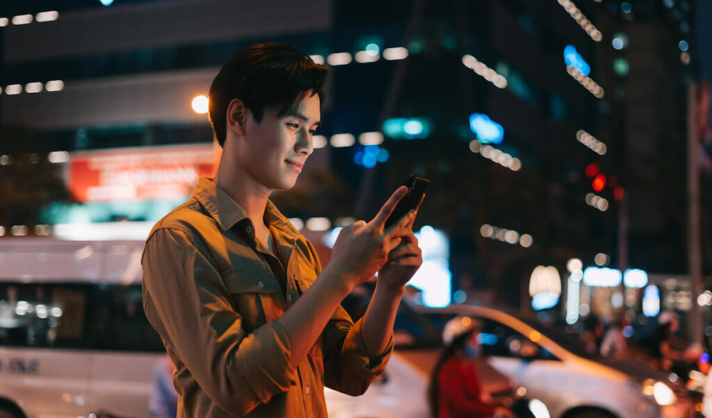 Young Asian man is using his phone while walking in the street at night