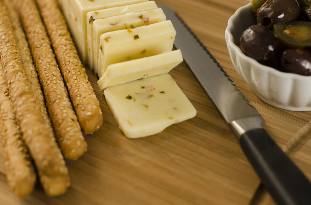 Pepper Jack cheese on a cutting board with some sesame seed bread sticks and a ramekin full of olives